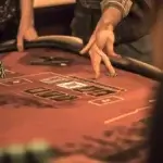 A close-up shot of a blackjack table with a player making a gesture, likely signaling their decision to the dealer. The player is seen pointing to the cards, indicating a possible action in the game. A stack of chips is visible on the table, with other players' hands and betting chips also in view, capturing the intensity of a casino gaming environment.