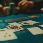 The image shows a close-up view of a blackjack table. The table is covered with a green felt surface, and several playing cards are spread out face-up. The visible cards include two aces and a king, suggesting that a game is in progress. In the background, there are a few stacks of poker chips, and the hands of players are slightly blurred, indicating movement. The scene is dimly lit, giving it an atmospheric and somewhat intense feel, typical of a casino environment.