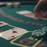 The image shows a close-up view of a blackjack table during a game. Two cards are laid face-up on the table—an Ace of Hearts and a King of Diamonds—indicating a blackjack hand. Nearby, several poker chips are stacked, with one prominently displaying a "$500" denomination. A player's hand is visible in the background, holding additional cards, possibly in the process of placing a bet. The table's green felt surface is marked with gold and red text, indicating different betting areas. The scene captures the high-stakes excitement and strategy involved in a game of blackjack.