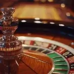The image shows a close-up of a roulette wheel in a casino. The focus is on the central spinning mechanism, which is highly polished and reflective, made of metal. Surrounding the spinner, the roulette wheel displays the numbers in alternating red and black sections, with a green section marked "0." The wood grain of the wheel is visible, adding a touch of elegance to the scene. The background is softly blurred, with warm lighting that enhances the luxurious and classic atmosphere of the casino setting.